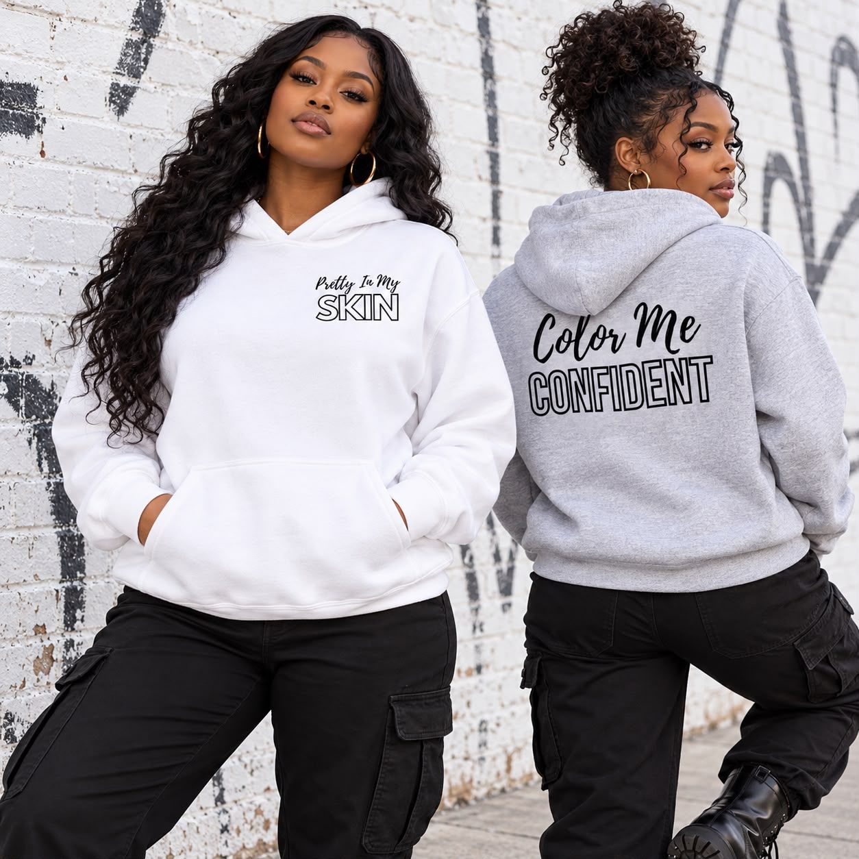 Two women in the white and ash grey Color Me Confident hoodies by a white brick wall