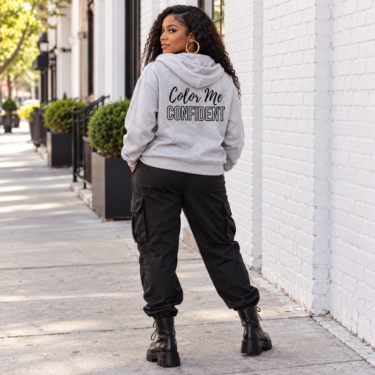 Woman in the ash grey Color Me Confident hoodie on a city sidewalk, back view